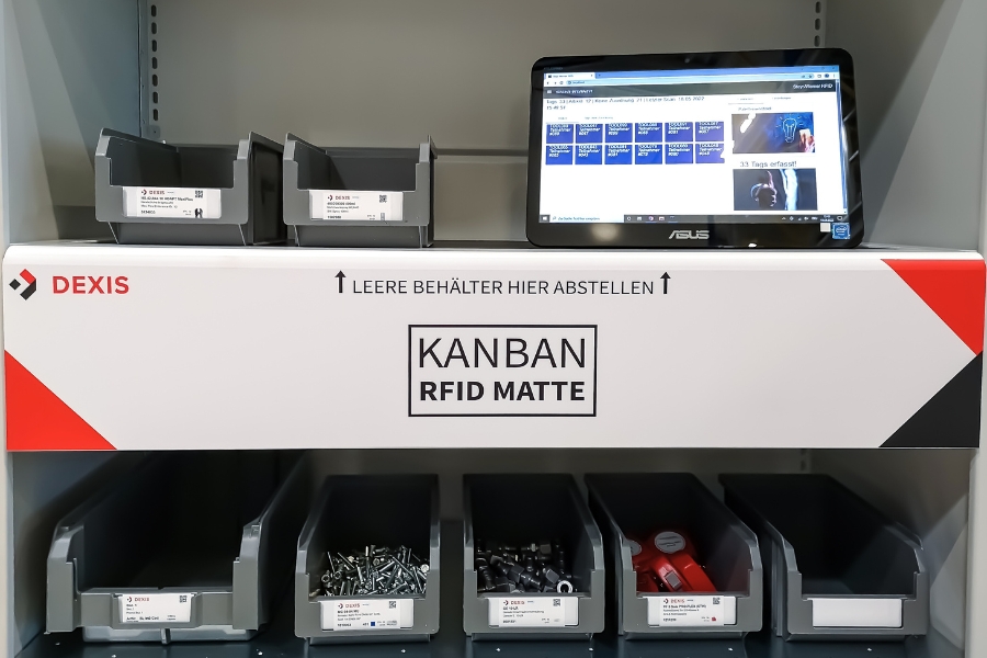 Das abgebildete Regal enthält sechs Behälter mit verschiedenen Kleinteilen. Ein leerer Behälterbereich ist durch einen Pfeil und den Text „LEERE BEHÄLTER HIER ABSTELLEN“ gekennzeichnet. Auf dem obersten Regal ist ein Monitor angebracht, auf dem eine Kanban-Bestandsverwaltungsschnittstelle angezeigt wird. Ein „DEXIS“-Logo ist sichtbar.